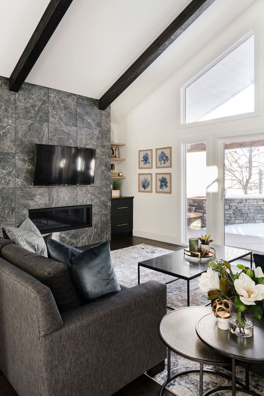 Modern living room featuring a dark gray sofa, a stone fireplace with a mounted television, and botanical wall art, complemented by natural light from skylights and a large window. A Calgary interior designer
