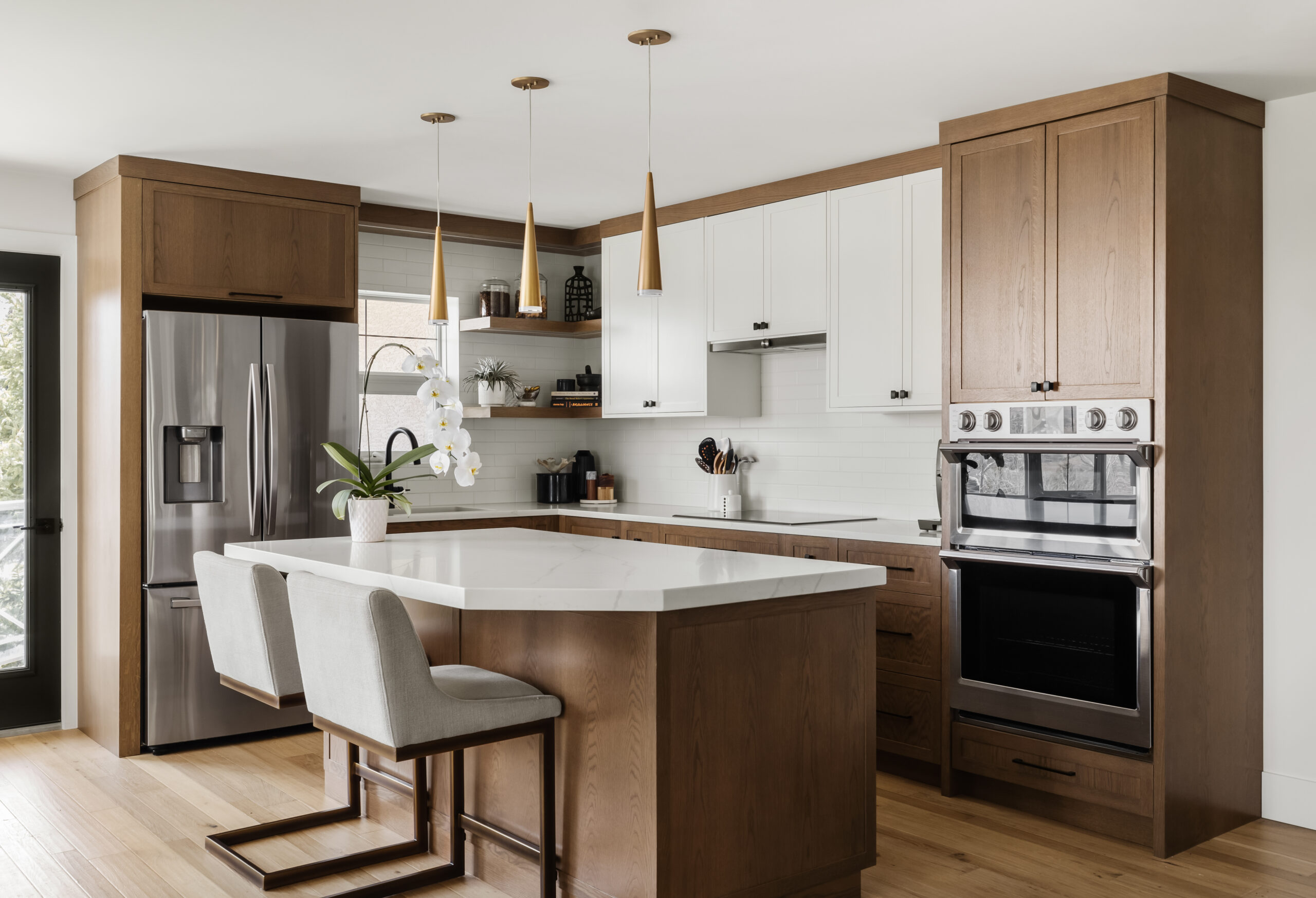 A modern kitchen features wooden cabinets, a stainless steel refrigerator, an oven, and a white marble island with two cushioned bar stools. Pendant lights hang above the island.