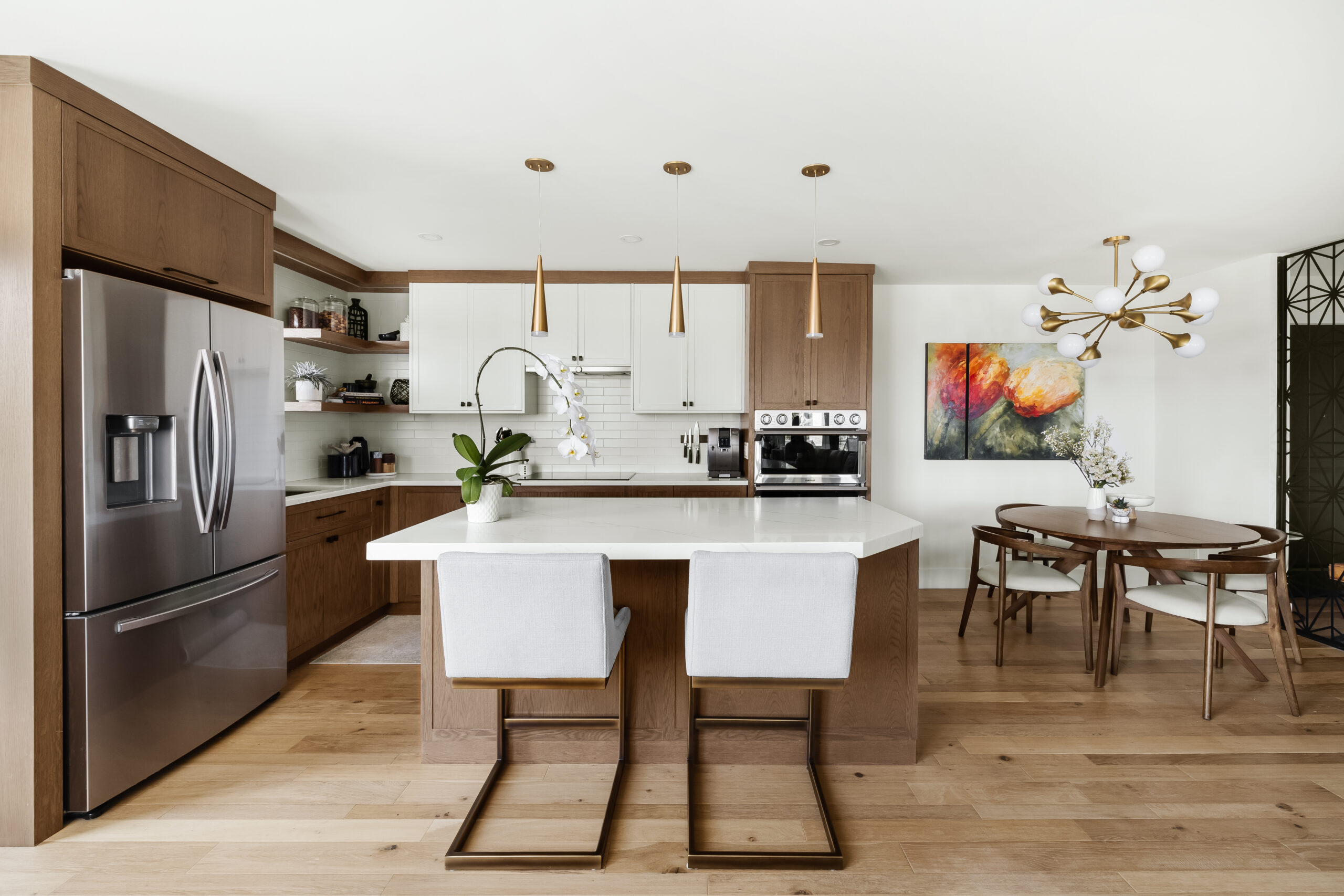 Modern kitchen with a central island, two chairs, stainless steel appliances, white countertops, wooden cabinets, and pendant lighting. A dining area with a table and chairs is visible in the background.