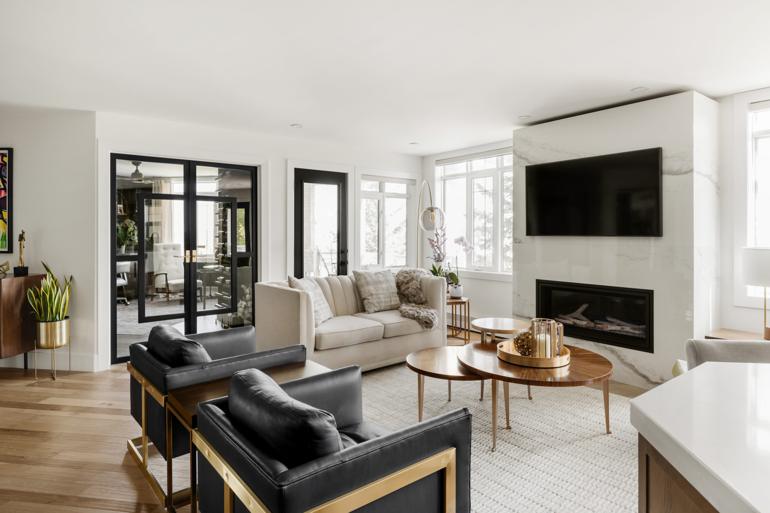 A modern living room with a light-colored sofa, black armchairs, coffee tables, a fireplace, and a wall-mounted TV. Large windows and glass doors provide natural light.