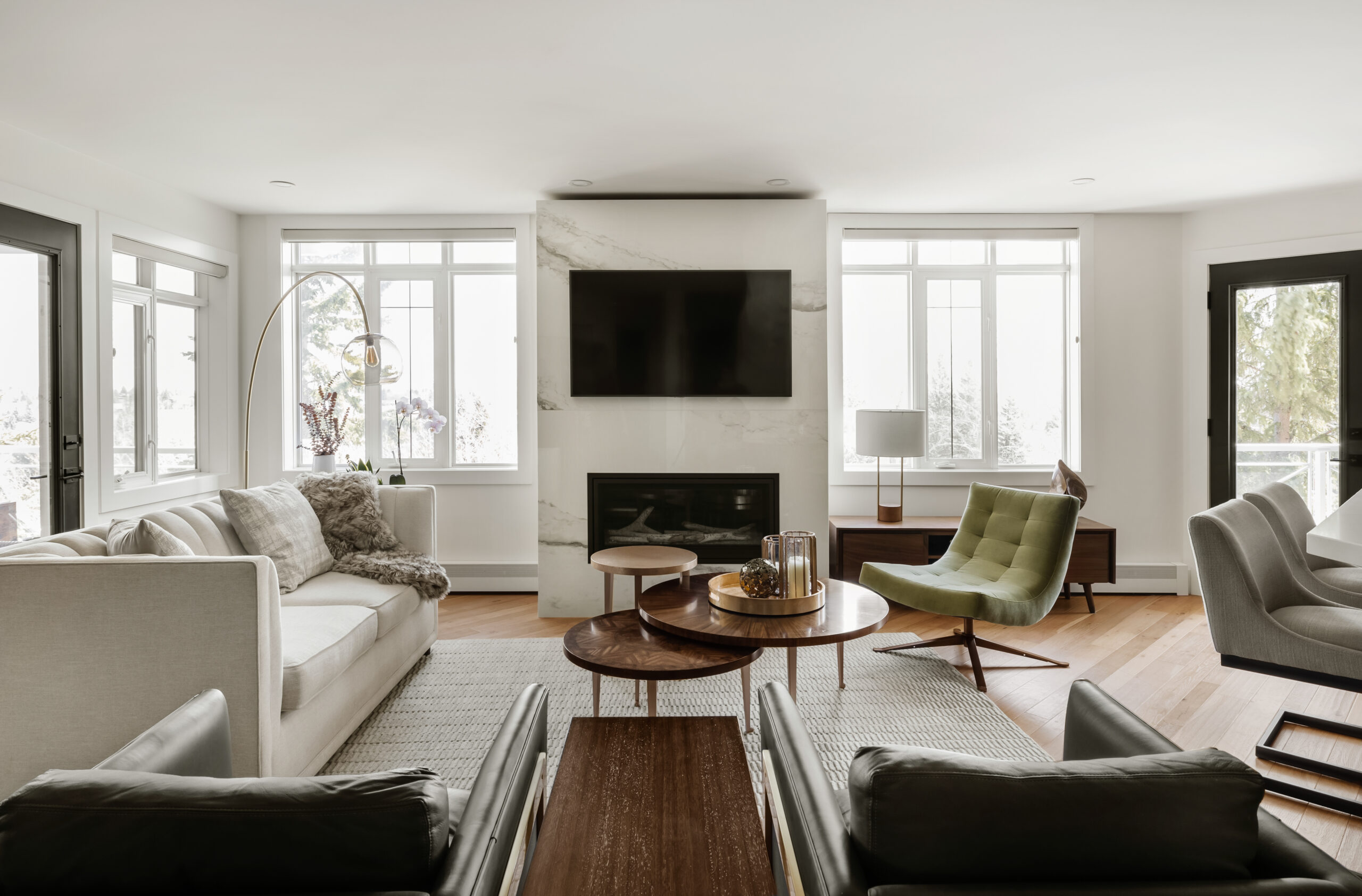 A modern living room with a light-colored sofa, black armchairs, coffee tables, a fireplace, and a wall-mounted TV. Large windows and glass doors provide natural light.