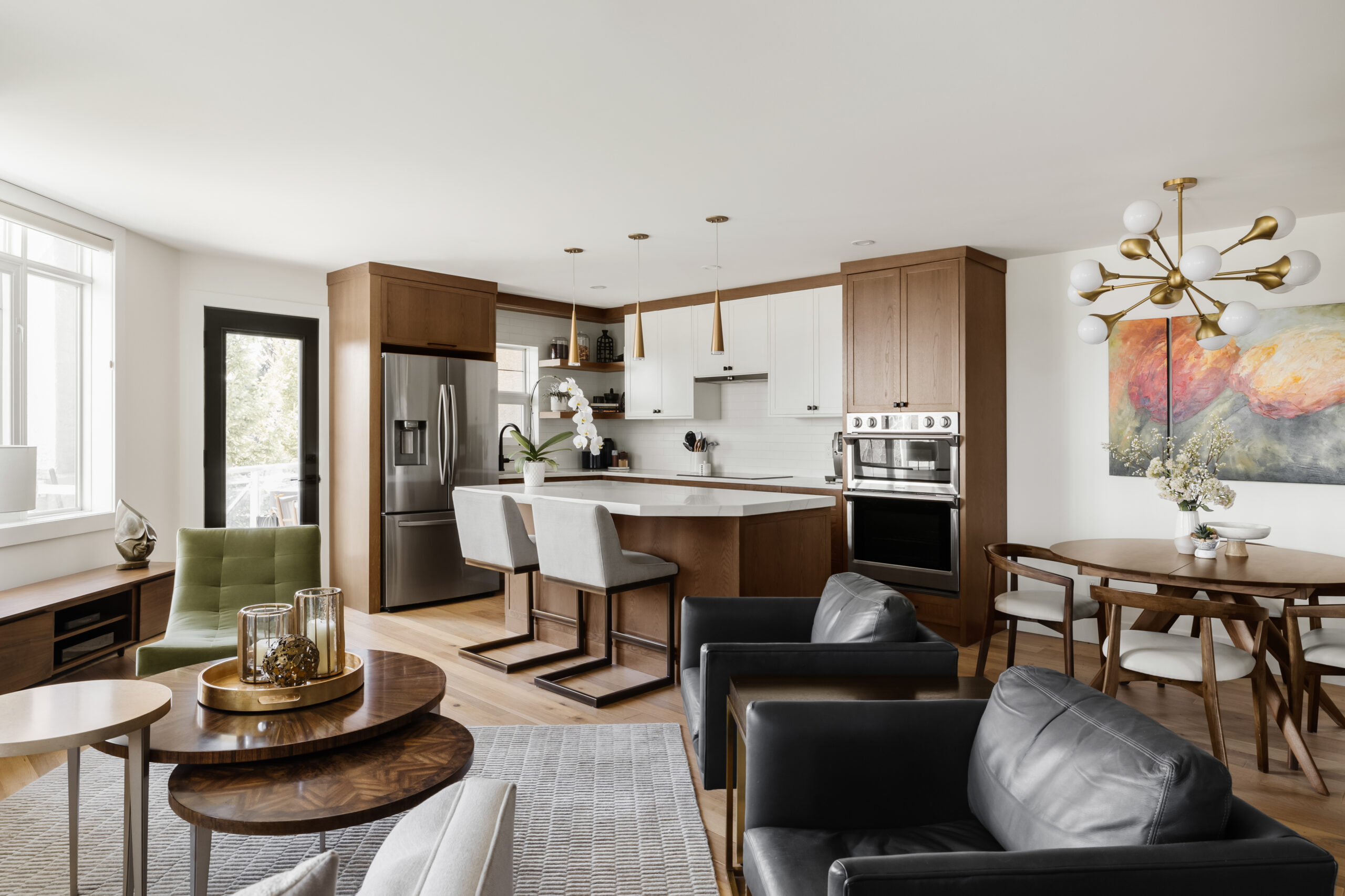 A modern kitchen features wooden cabinets, a stainless steel refrigerator, an oven, and a white marble island with two cushioned bar stools. Pendant lights hang above the island.