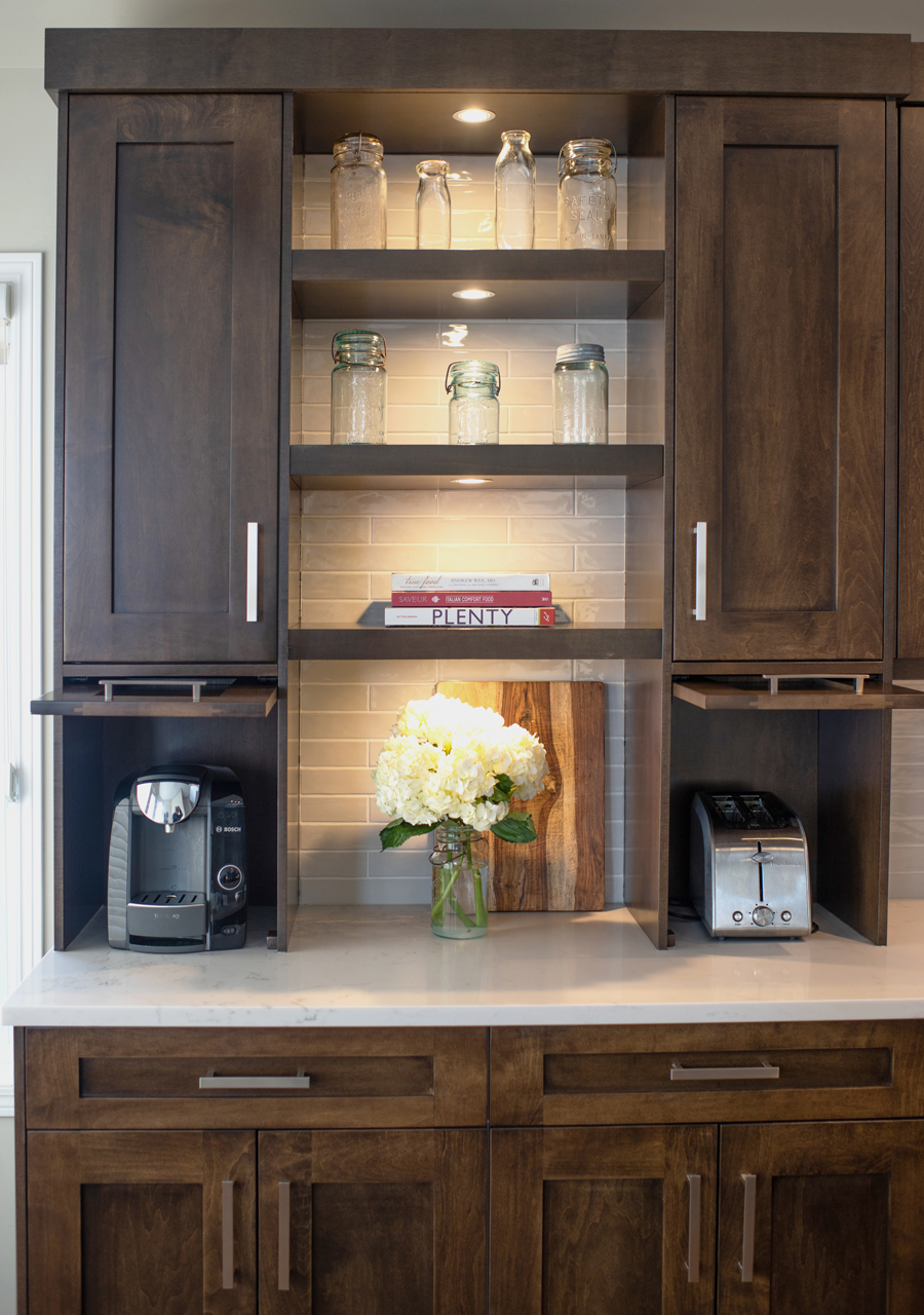 A kitchen counter with dark wooden cabinets, open shelves displaying mason jars, books, and a vase of white flowers, alongside a coffee maker, a cutting board, and a toaster.