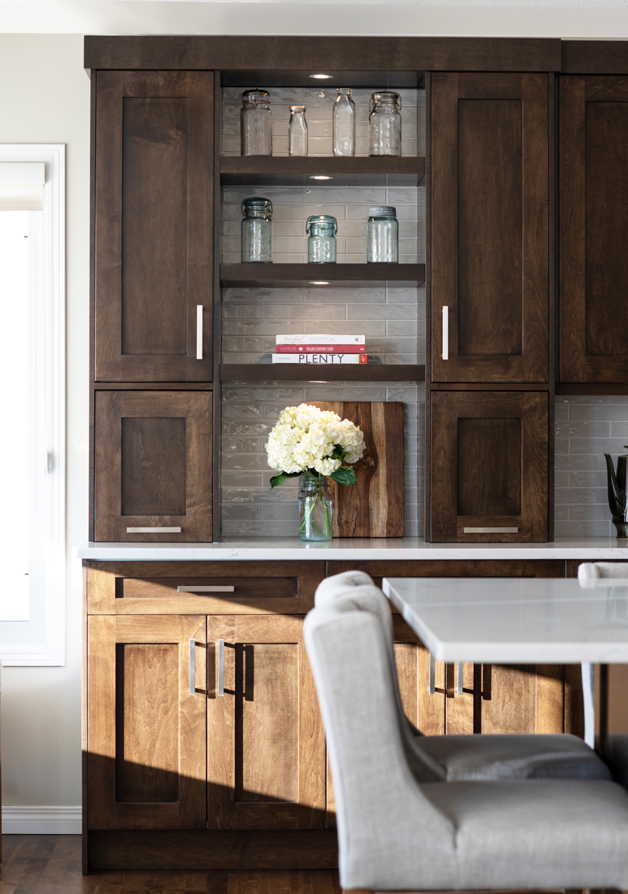 A kitchen scene featuring dark wooden cabinets, glass jars on open shelves, a vase of white flowers, and a light grey upholstered chair.
