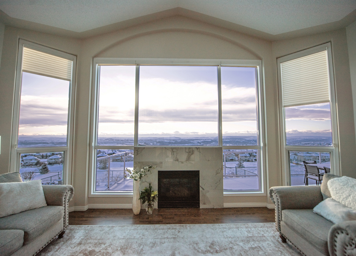 A living room with a large window offering a panoramic view of a snowy landscape, with white walls, two armchairs, a fireplace, and a vase of flowers on the mantel.