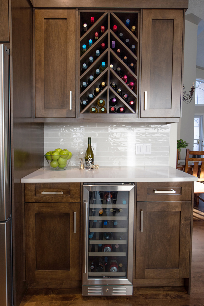 A kitchen area featuring a wine fridge, wine storage, wooden cabinets, a white countertop with a bottle of wine and apples, and a white tile backsplash.