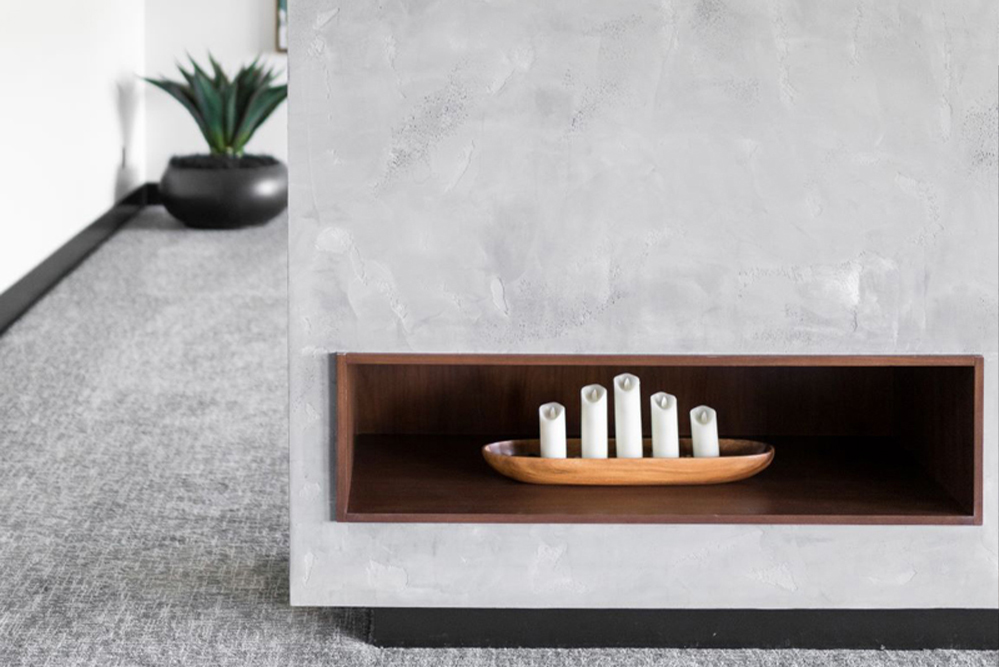 Concrete wall with a built-in wooden shelf holding a wooden tray with five white cylindrical objects, and a plant in a black pot in the background.