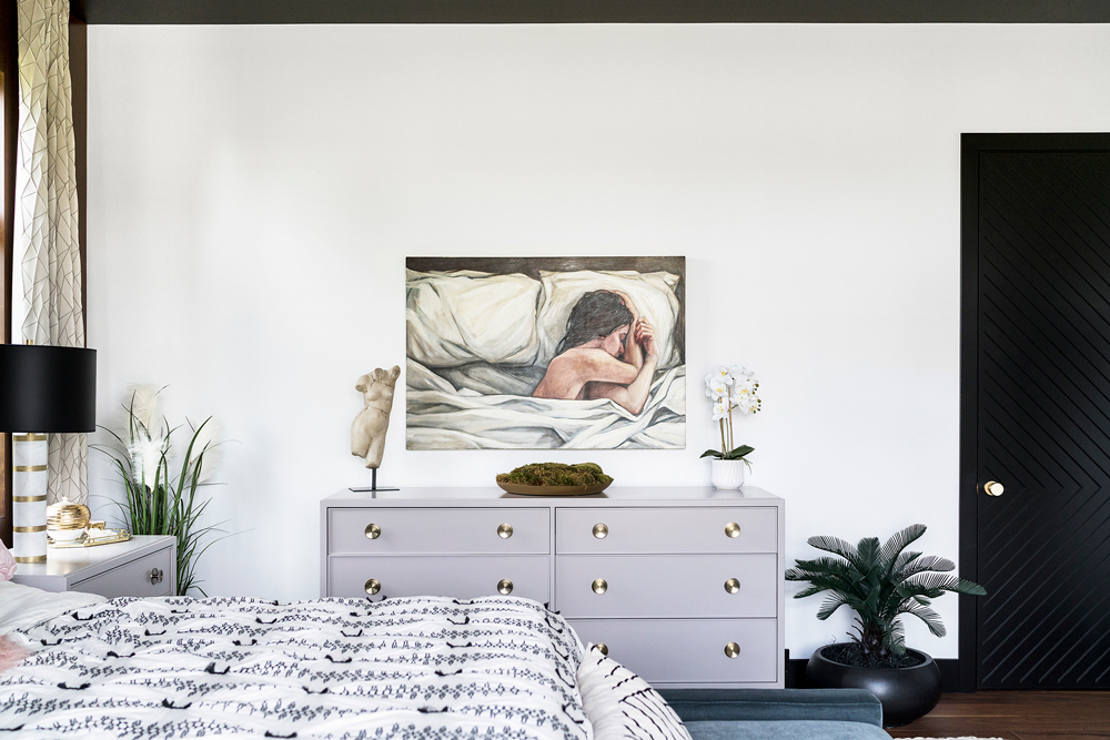 A bedroom with a white dresser against a white wall, featuring a painting of a person resting. The room includes a bed with patterned bedding, a black lamp, plants, and a door with a diagonal pattern.