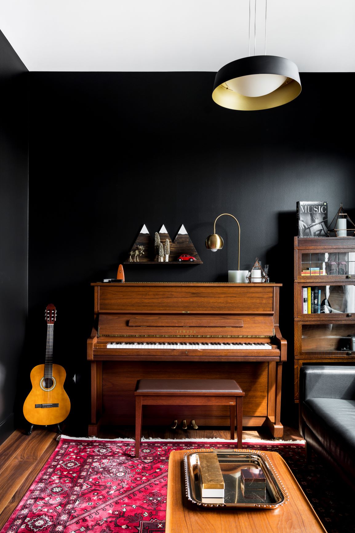 A seductive music room with a piano, guitar, bookshelf, and a red patterned rug. A coffee table with a tray is in the foreground, and a modern pendant light hangs from the ceiling.