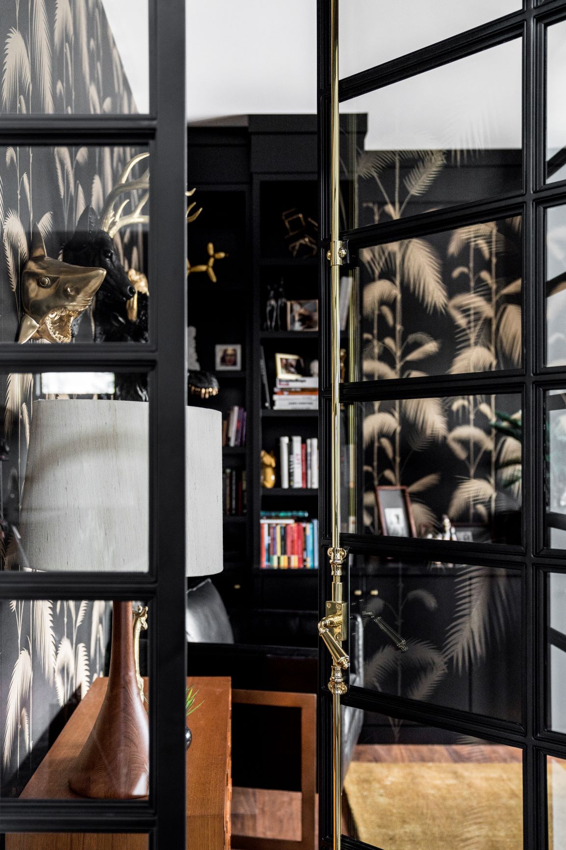 A modern home office with black-framed glass doors, tropical-themed wallpaper, a desk with a white lamp, and a bookshelf filled with books in the background.