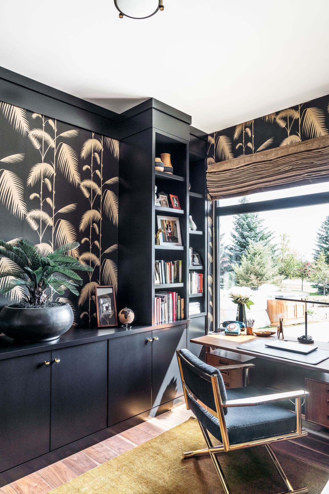 A modern home office features a black and gold palm-leaf wall, built-in shelving with books and decor, a window with a nature view, a wooden desk, and a black chair with metal accents.