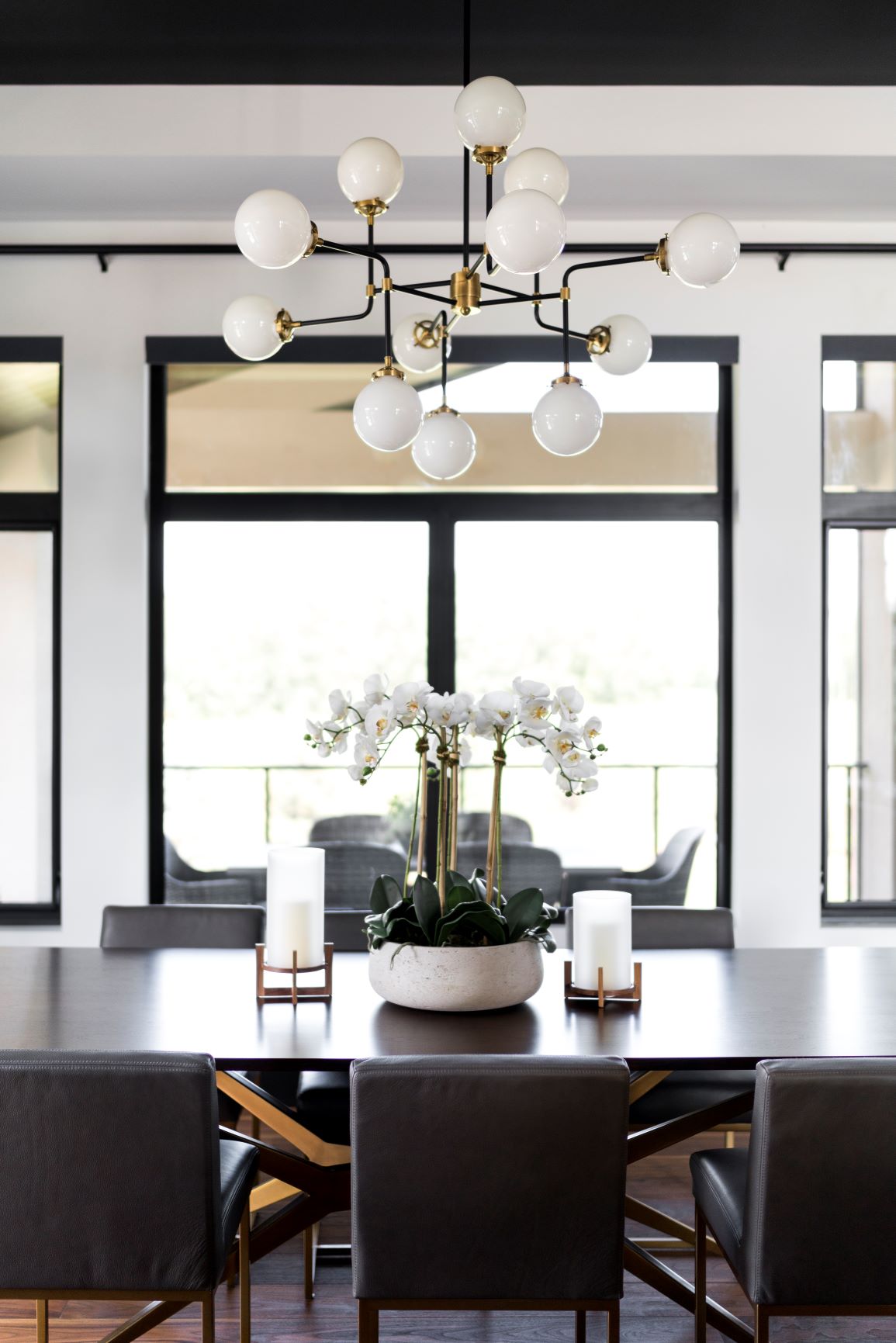 Modern dining room with a dark wooden table, black chairs, white orchid centerpiece, two candles, and a contemporary chandelier. 