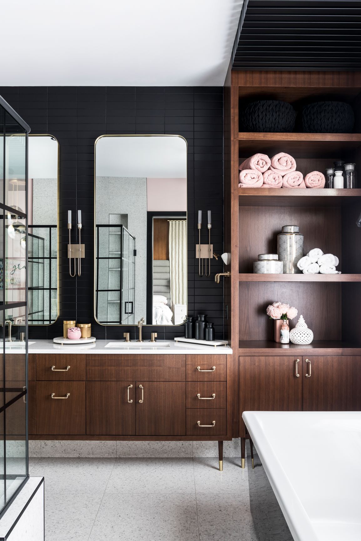 Modern bathroom with dark wall tiles, wooden cabinets, double mirrors, and open shelving stocked with towels and toiletries. A glass shower and white bathtub are also visible.