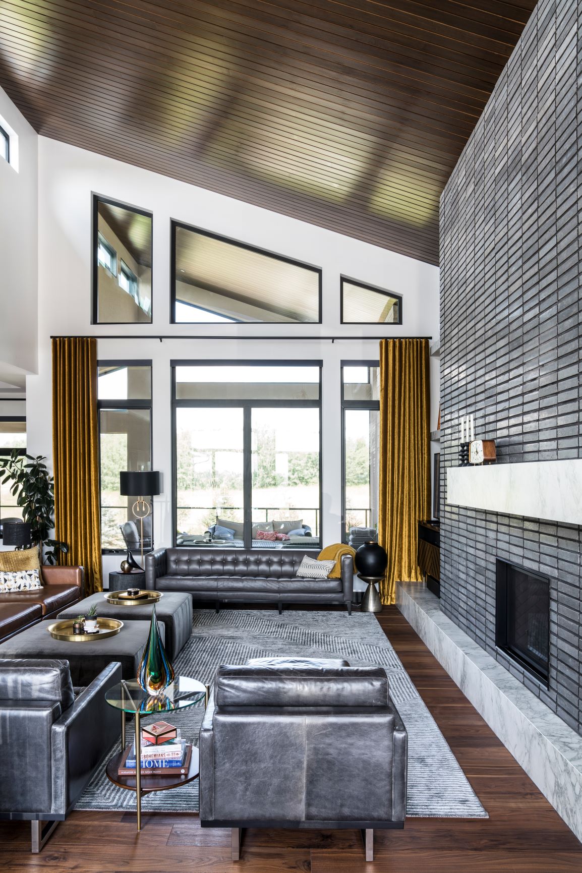 A modern living room with gray sofas, a large fireplace, tall windows with mustard curtains, and a vaulted wooden ceiling. Accessories include a rug, lamps, and books on a side table.