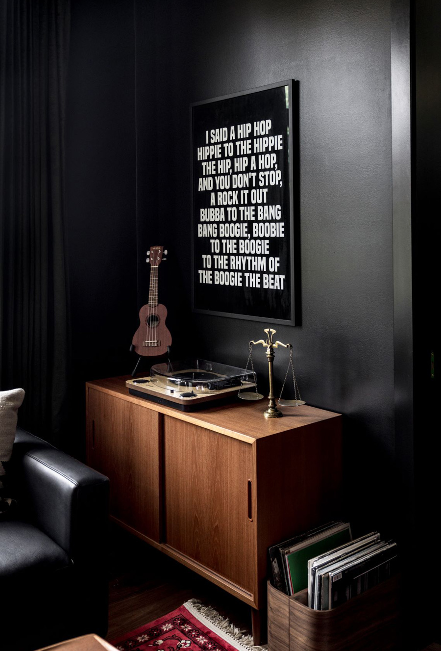 A wooden sideboard with a record player and a small guitar against a dark wall. A framed poster with hip hop lyrics by The Sugarhill Gang hangs above the sideboard.