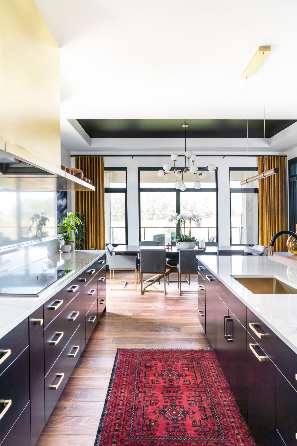 Modern kitchen with black cabinets, white countertops, a red rug, gold handles, and a view into a dining area.