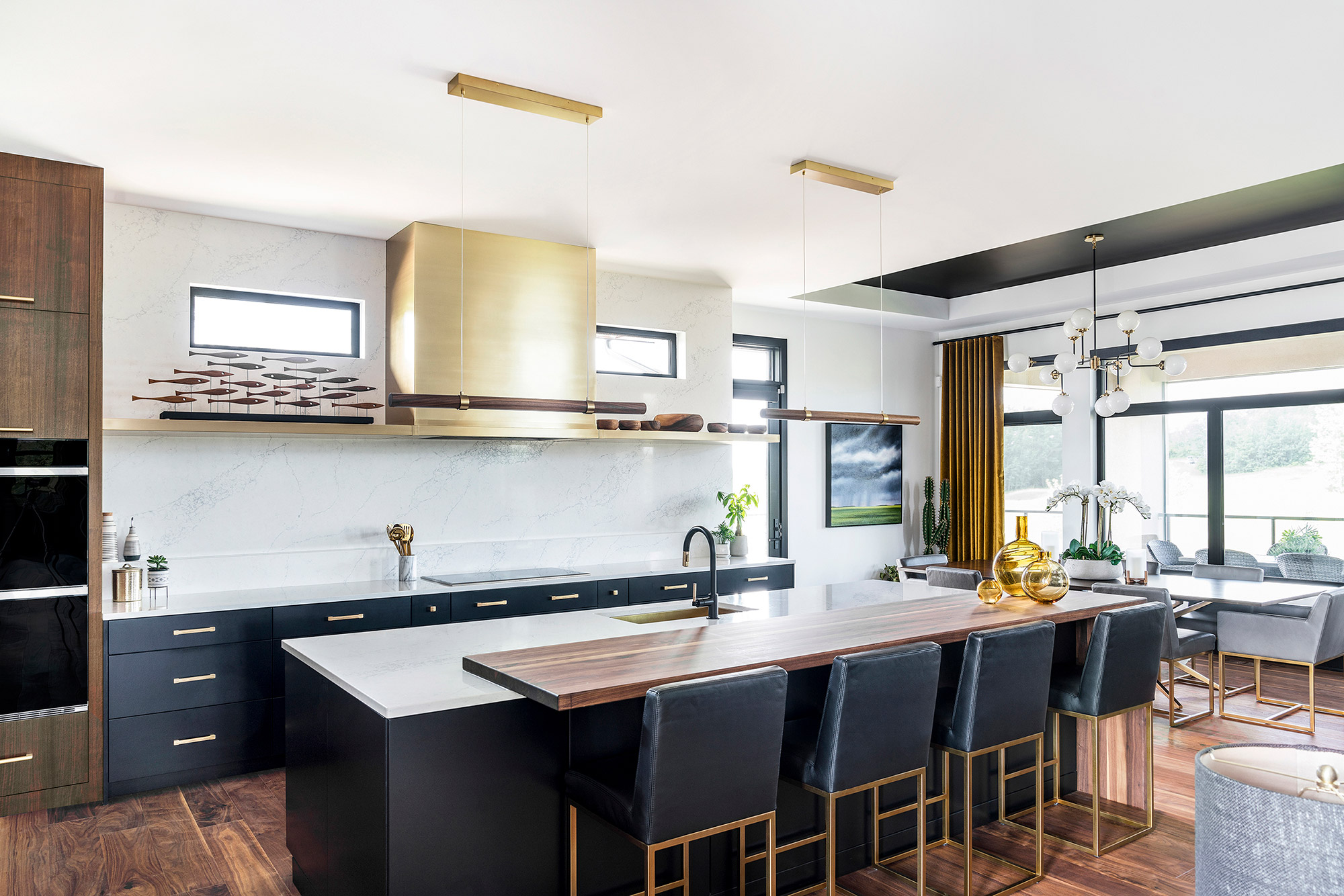 Modern kitchen with dark cabinets, a large wooden island, brass fixtures, and a built-in wine rack. Dining area in the background with a panoramic window view.