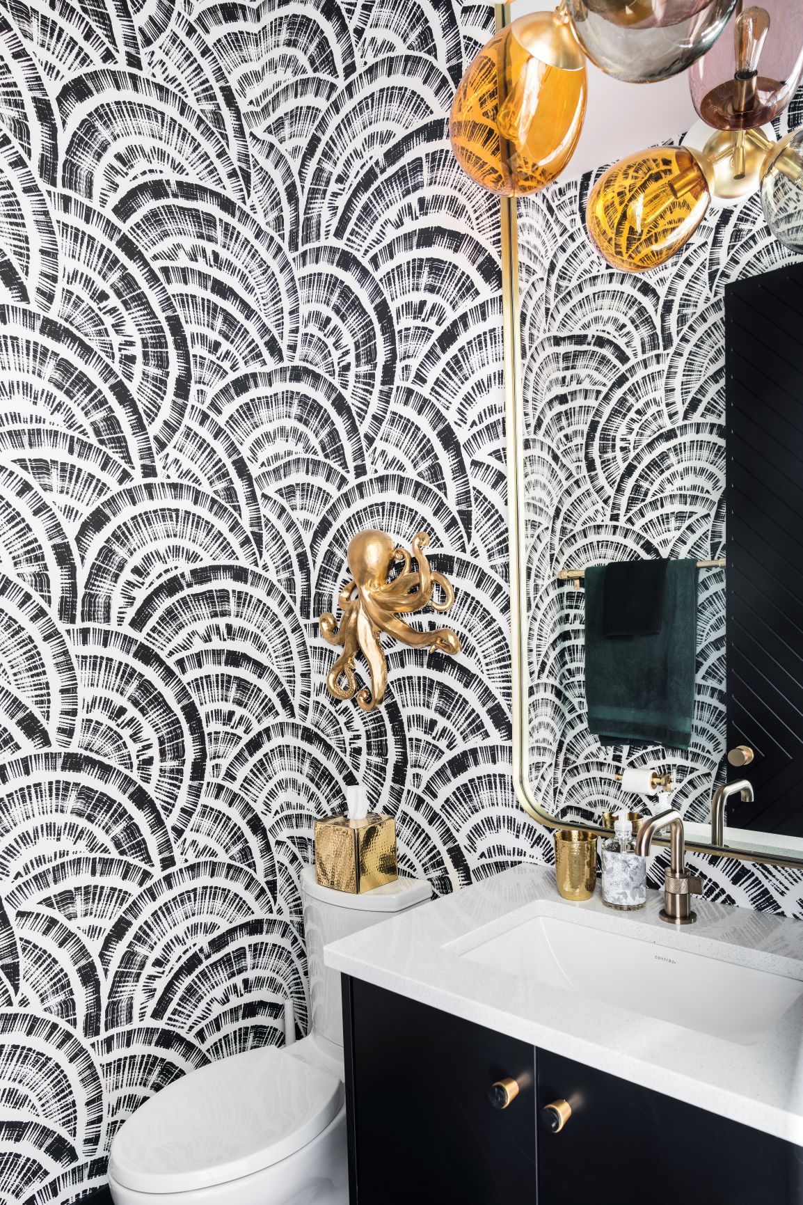 A modern bathroom with black and white patterned wallpaper, gold fixtures, a white sink, black vanity, round mirror.