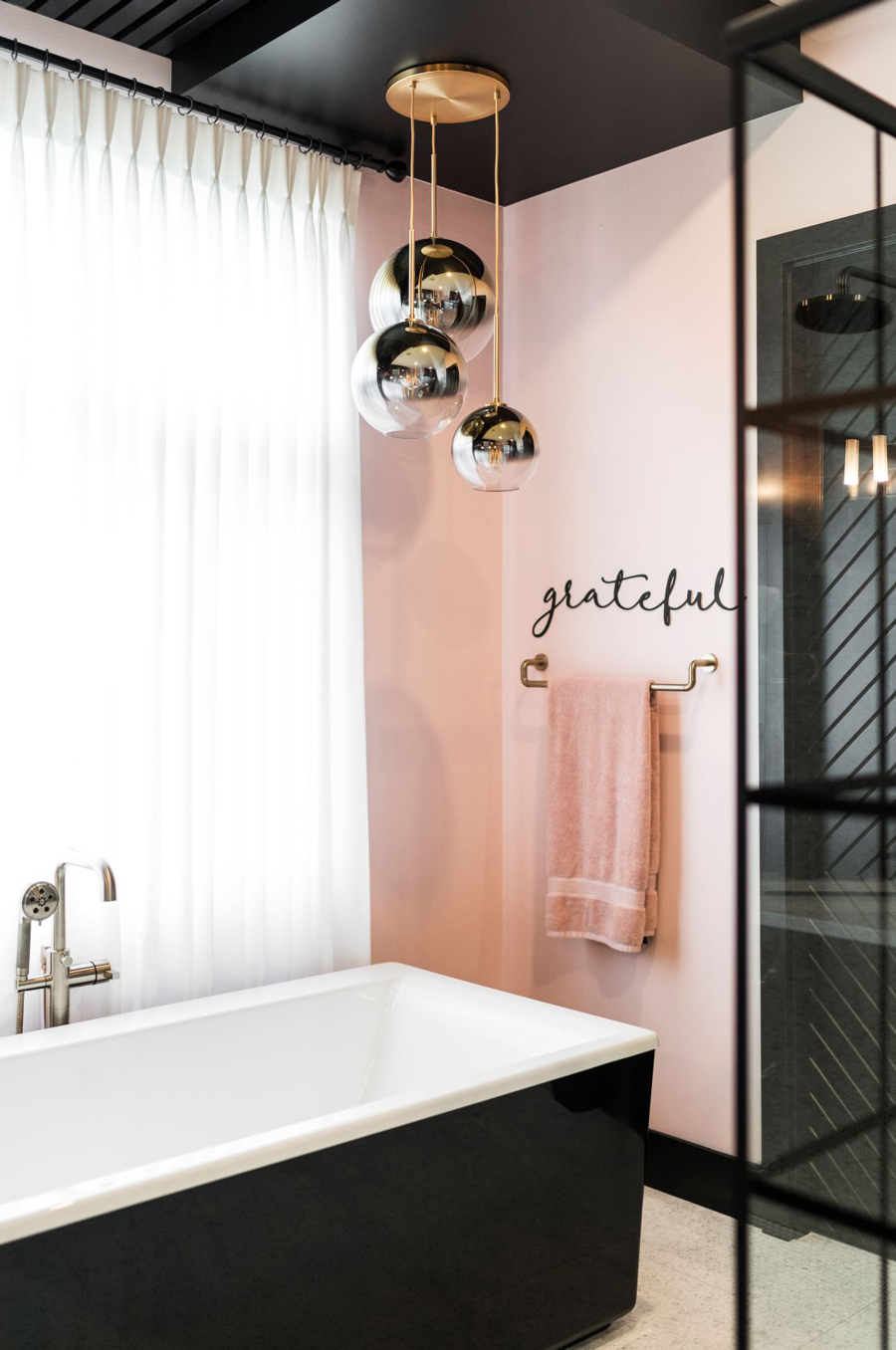 A modern bathroom features a black and white bathtub, pink wall with a "grateful" sign, hanging metallic spherical lights, and a pink towel on a gold towel rack.