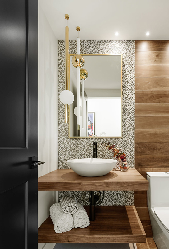 Modern bathroom with a wooden vanity, white vessel sink, round mirror, hanging pendant lights, and open shelving with rolled towels and decorative branches. Décor includes wood and mosaic tile accents.