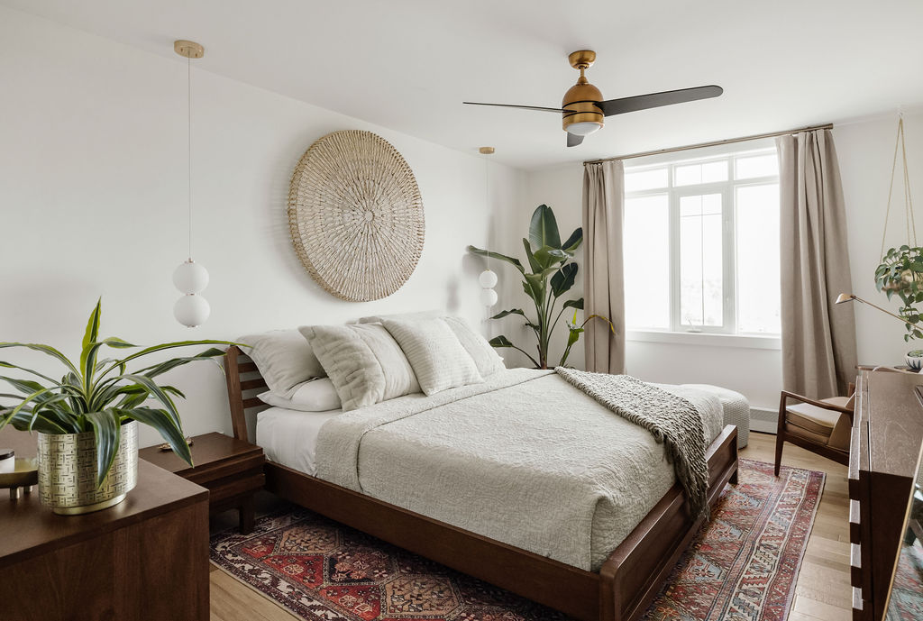 A bright bedroom with a large bed, plants, a decorative wall piece, a ceiling fan, and a window with light curtains. The floor has a patterned rug under the bed. Two hanging lamps are beside the bed.