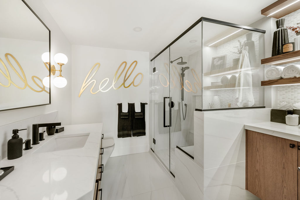 Modern bathroom with a glass-enclosed shower, large mirror, lighted vanity, and minimalist decor. "Hello" is written in large gold script on the wall above black towels.