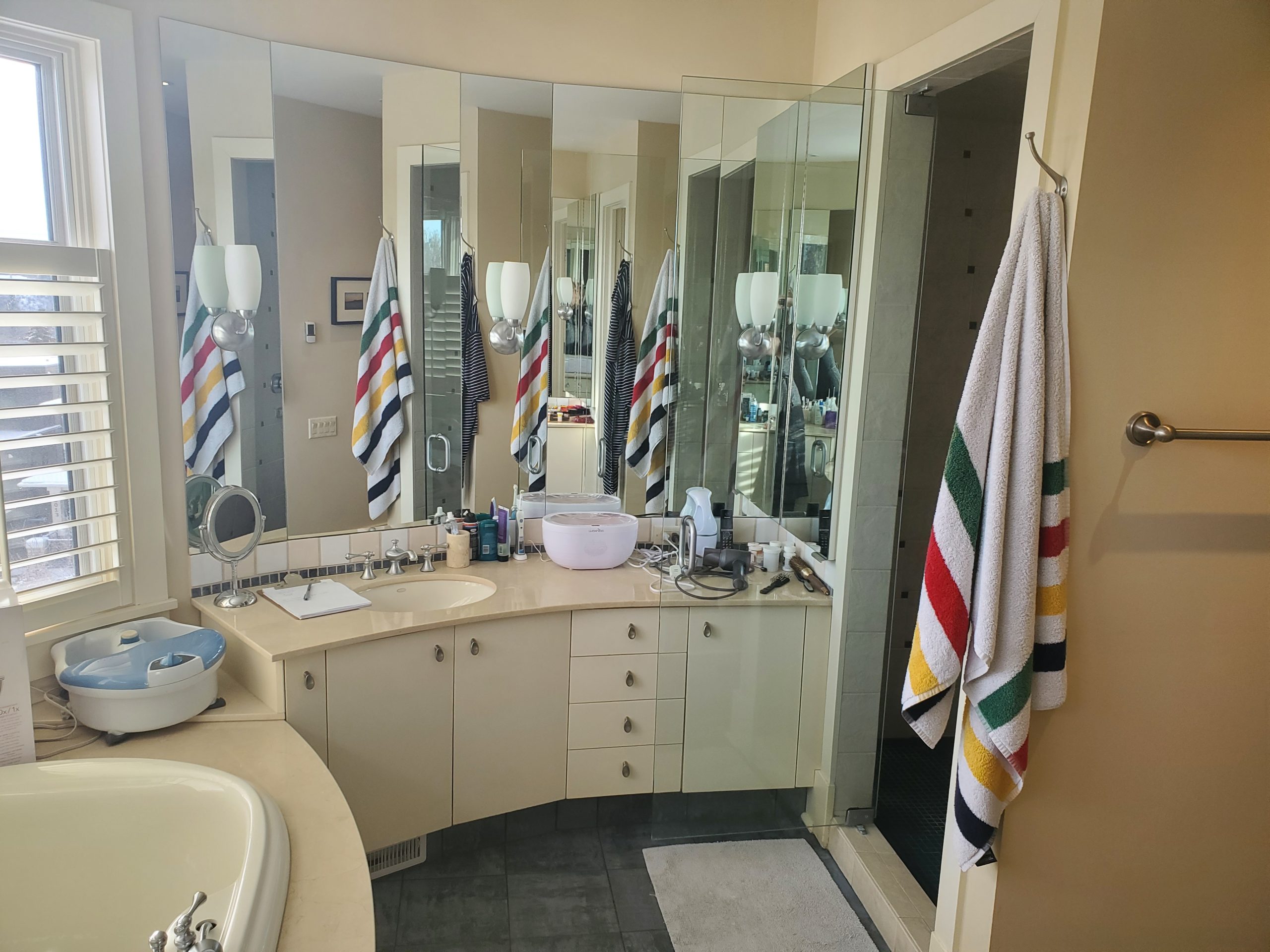 StudioRuInterior-Design_Calgary_Mount-Royal_Bathroom-Before_Vanity This image showcases a bathroom before renovation, featuring a glass-enclosed shower with dark tile flooring and a mix of square and rectangular wall tiles. The towel with colorful stripes adds a playful contrast to the otherwise neutral tones of the space. The vanity area, visible on the left, has a cluttered countertop, while the background reveals a walk-in closet with open shelving. The design elements, including warm beige walls and classic lighting, give the space a dated yet functional appearance, making it a strong candidate for a modern refresh.