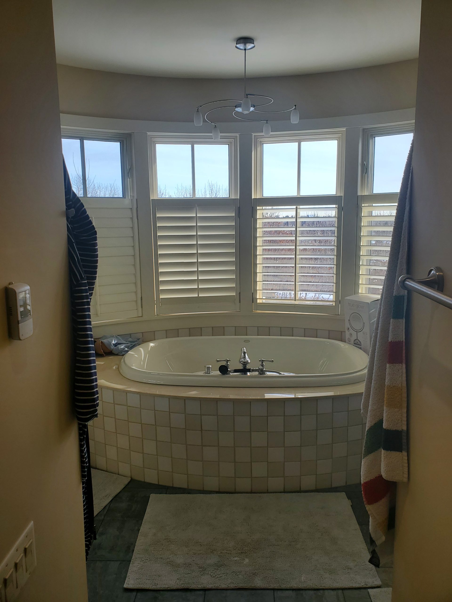 StudioRuInterior-Design_Calgary_Mount-Royal_Bathroom-Before_tub This image showcases a bathroom before renovation, featuring a glass-enclosed shower with dark tile flooring and a mix of square and rectangular wall tiles. The towel with colorful stripes adds a playful contrast to the otherwise neutral tones of the space. The vanity area, visible on the left, has a cluttered countertop, while the background reveals a walk-in closet with open shelving. The design elements, including warm beige walls and classic lighting, give the space a dated yet functional appearance, making it a strong candidate for a modern refresh.