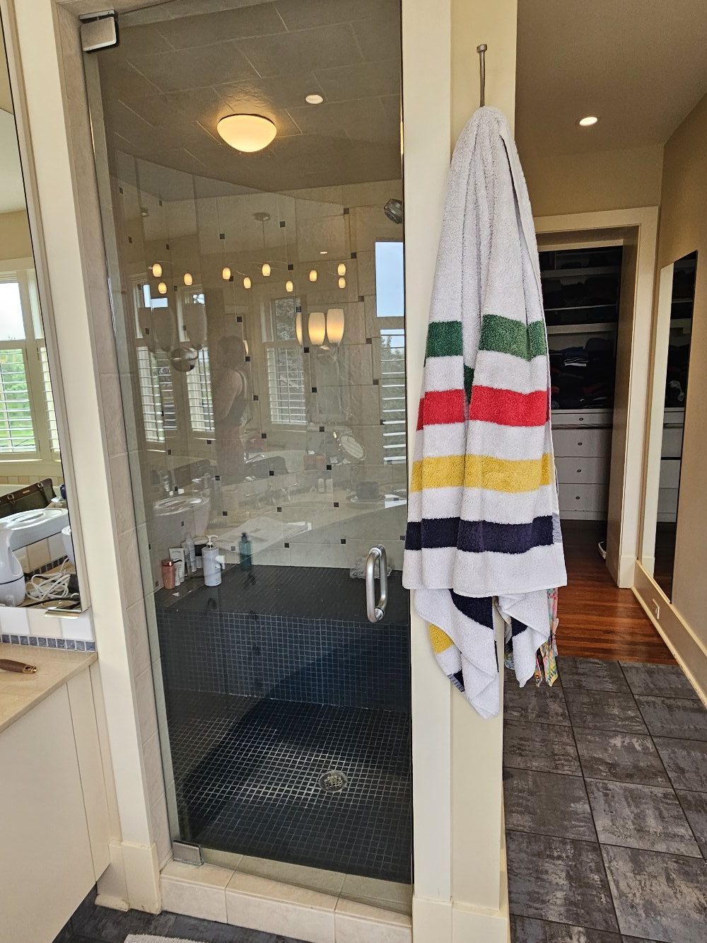 StudioRuInterior-Design_Calgary_Mount-Royal_Bathroom-Before_Shower This image showcases a bathroom before renovation, featuring a glass-enclosed shower with dark tile flooring and a mix of square and rectangular wall tiles. The towel with colorful stripes adds a playful contrast to the otherwise neutral tones of the space. The vanity area, visible on the left, has a cluttered countertop, while the background reveals a walk-in closet with open shelving. The design elements, including warm beige walls and classic lighting, give the space a dated yet functional appearance, making it a strong candidate for a modern refresh.