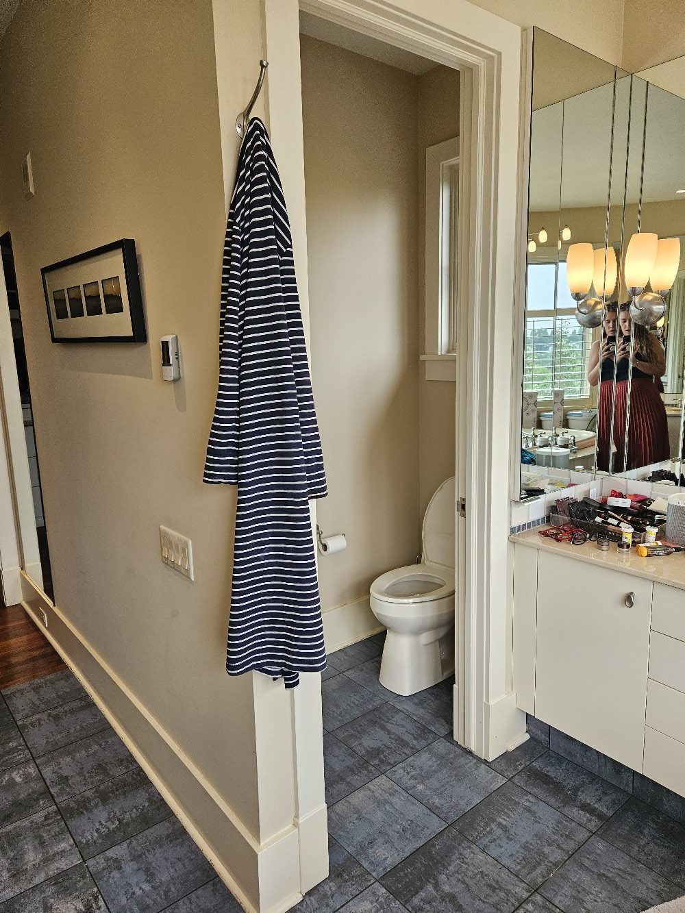 StudioRuInterior-Design_Calgary_Mount-Royal_Bathroom-Before_Bathroom This image showcases a bathroom before renovation, featuring a glass-enclosed shower with dark tile flooring and a mix of square and rectangular wall tiles. The towel with colorful stripes adds a playful contrast to the otherwise neutral tones of the space. The vanity area, visible on the left, has a cluttered countertop, while the background reveals a walk-in closet with open shelving. The design elements, including warm beige walls and classic lighting, give the space a dated yet functional appearance, making it a strong candidate for a modern refresh.