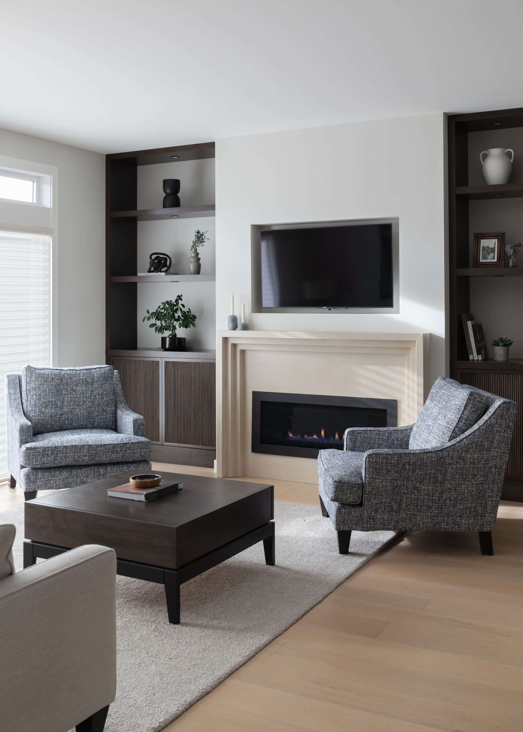 Modern living room with two gray armchairs facing a wooden coffee table. A television is mounted above a fireplace. Shelves with decorative items flank the fireplace. Light wood flooring.