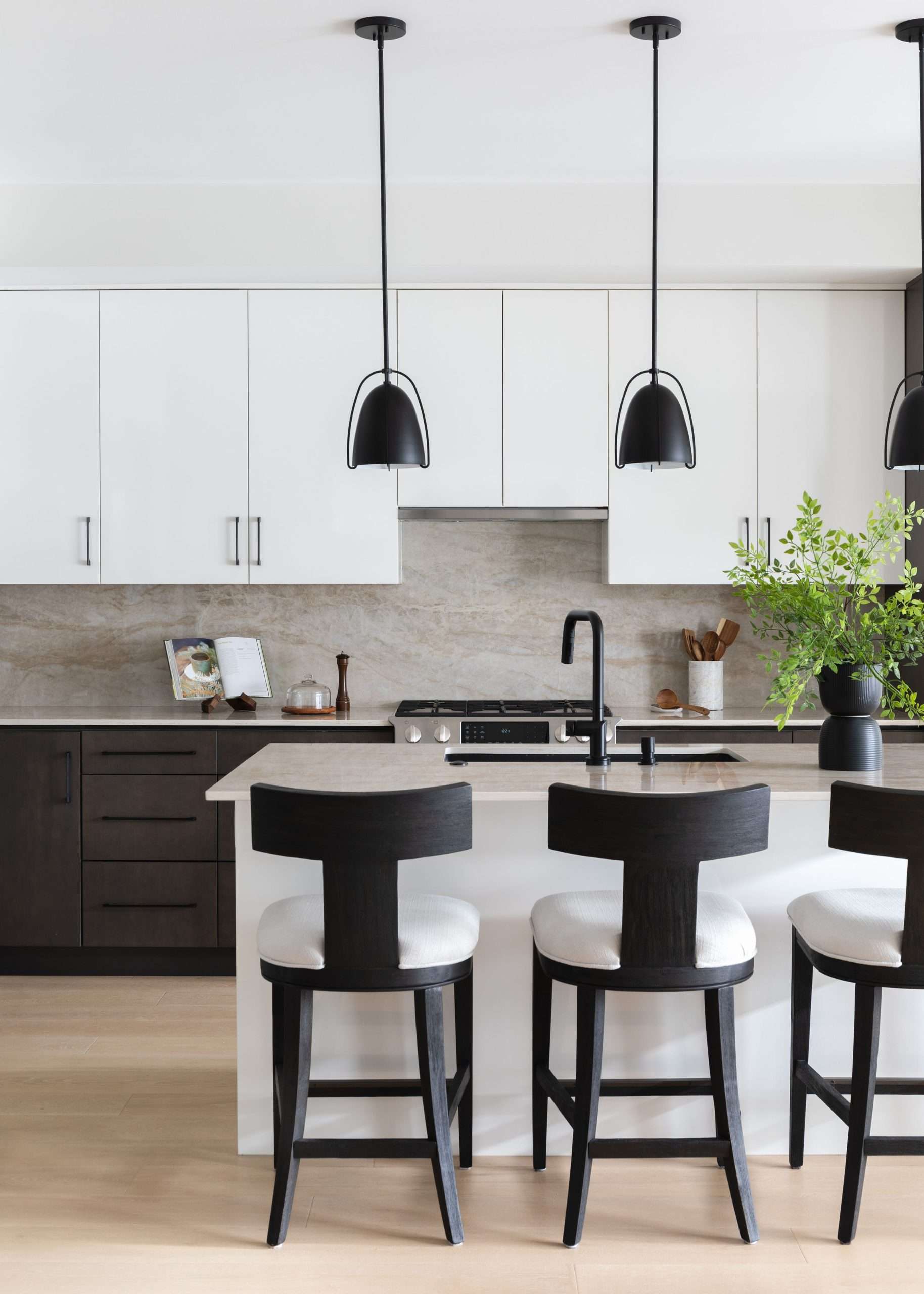 Modern kitchen with white cabinets, black pendant lights, and a central island featuring black and white bar stools. A vase with green foliage is on the counter.