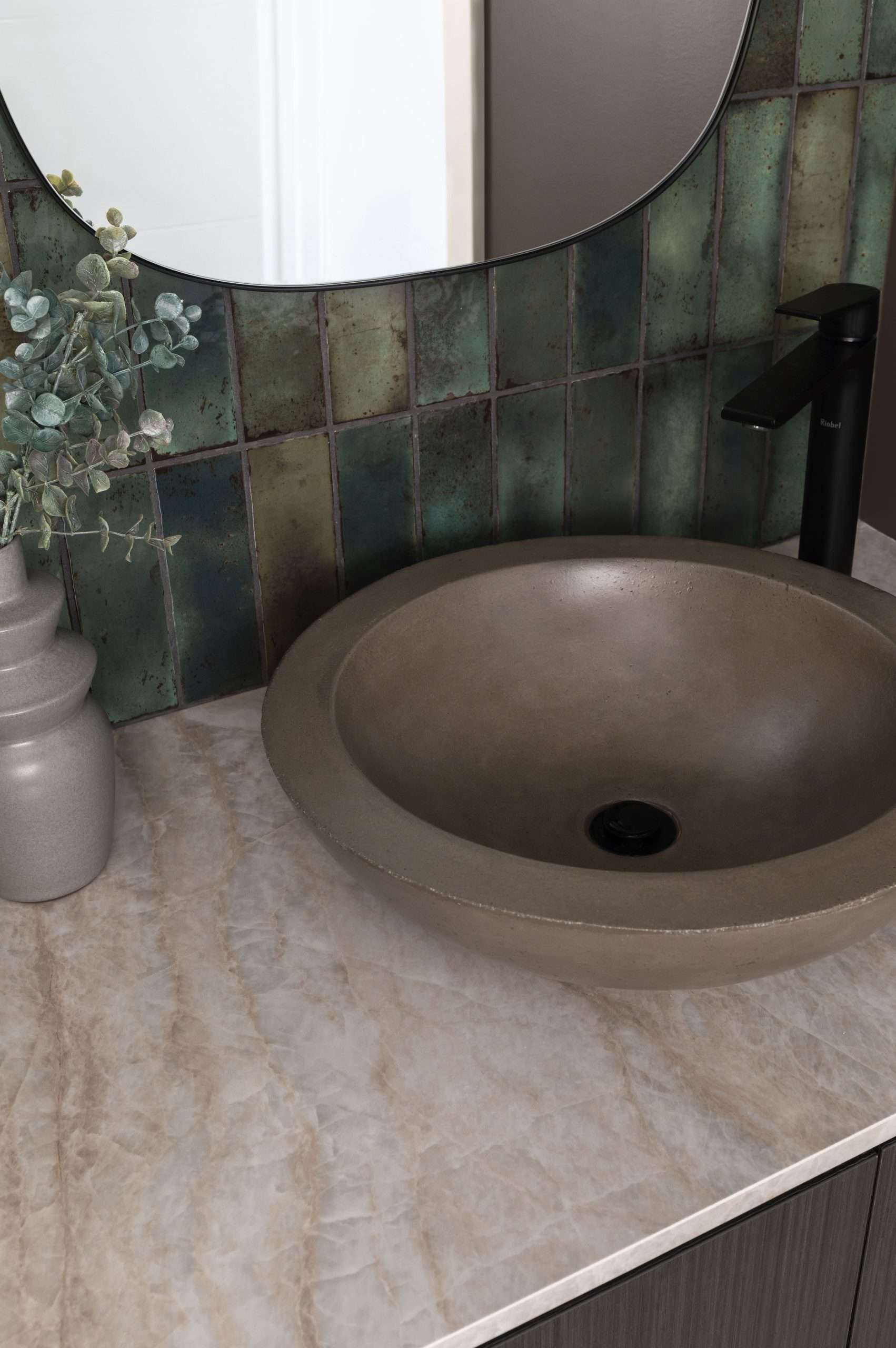 A modern bathroom vanity featuring a round, stone-like vessel sink with a matte finish, a sleek black faucet, and a beige marble-look countertop. The backsplash consists of rustic green-toned square tiles with an aged, weathered appearance. A curved mirror with a thin black frame is partially visible, reflecting a bright doorway. A decorative gray vase with dried eucalyptus adds a natural touch to the design.