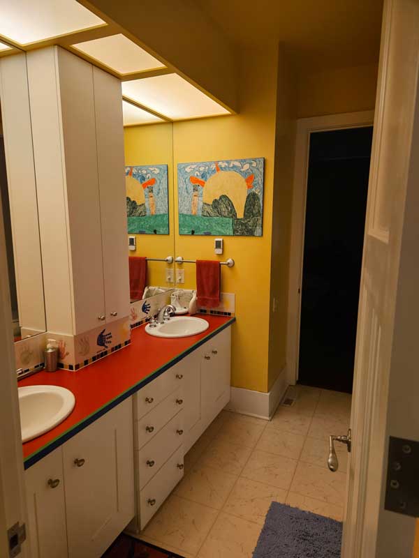 StudioRuInterior-Design_Calgary_Mount-Royal_Bathroom-Before_Shared_Bath A bathroom with yellow walls and a bold red countertop before renovation. The space features double sinks, white cabinetry, and large mirrors. The countertop is cluttered with personal care items, a hairbrush, and a toiletry bag. Red hand towels and playful artwork with a colorful animal design decorate the walls. The lighting consists of recessed ceiling panels, and the floor has a Spider-Man-themed rug. The overall look is vibrant but somewhat dated and in need of an update.