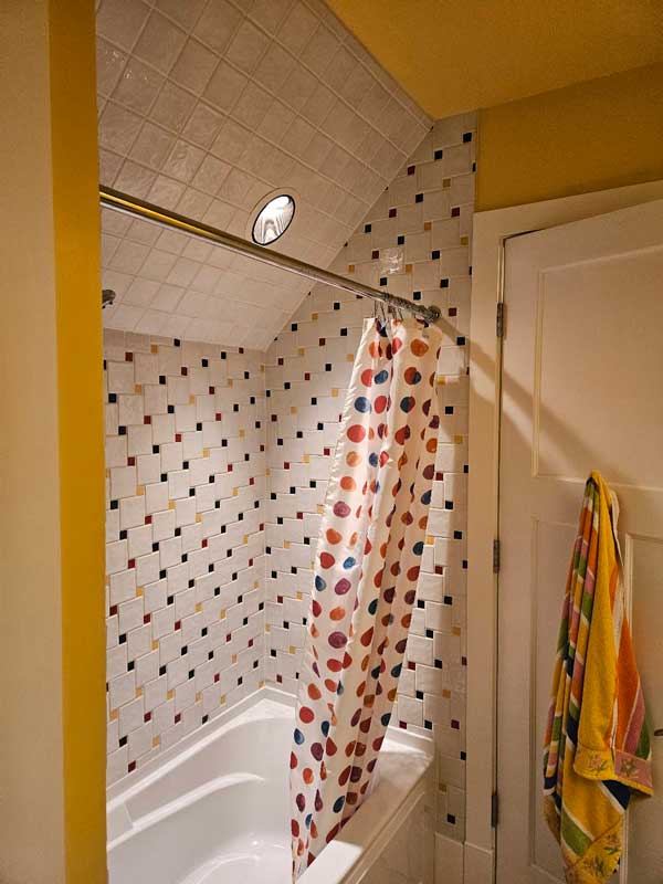 StudioRuInterior-Design_Calgary_Mount-Royal_Bathroom-Before_Shared_Bath_shower A bathroom with yellow walls and a bold red countertop before renovation. The space features double sinks, white cabinetry, and large mirrors. The countertop is cluttered with personal care items, a hairbrush, and a toiletry bag. Red hand towels and playful artwork with a colorful animal design decorate the walls. The lighting consists of recessed ceiling panels, and the floor has a Spider-Man-themed rug. The overall look is vibrant but somewhat dated and in need of an update.