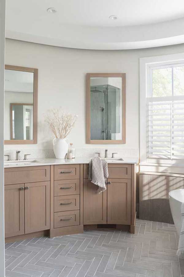StudioRuInterior-Design_Calgary_Mount-Royal_Bathroom-Curved-Vanity This bathroom exudes elegance and relaxation with its freestanding soaking tub placed in front of a curved wall of large windows, fitted with white plantation shutters that allow natural light to filter through. The combination of neutral tones, herringbone-patterned flooring, and textured wall paneling adds depth and sophistication. A wooden stool holding a potted green plant and a candle adds warmth, while draped towels over the tub enhance the inviting, spa-like atmosphere. The sleek floor-mounted faucet completes the modern yet timeless aesthetic.