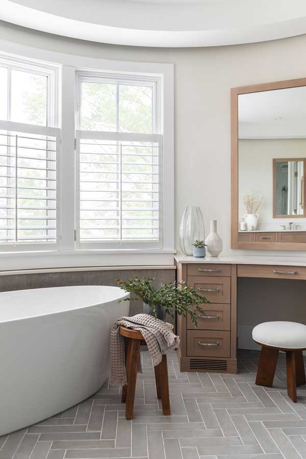 StudioRuInterior-Design_Calgary_Mount-Royal_Bathroom-Curved_Makeupdesk This bathroom exudes elegance and relaxation with its freestanding soaking tub placed in front of a curved wall of large windows, fitted with white plantation shutters that allow natural light to filter through. The combination of neutral tones, herringbone-patterned flooring, and textured wall paneling adds depth and sophistication. A wooden stool holding a potted green plant and a candle adds warmth, while draped towels over the tub enhance the inviting, spa-like atmosphere. The sleek floor-mounted faucet completes the modern yet timeless aesthetic.