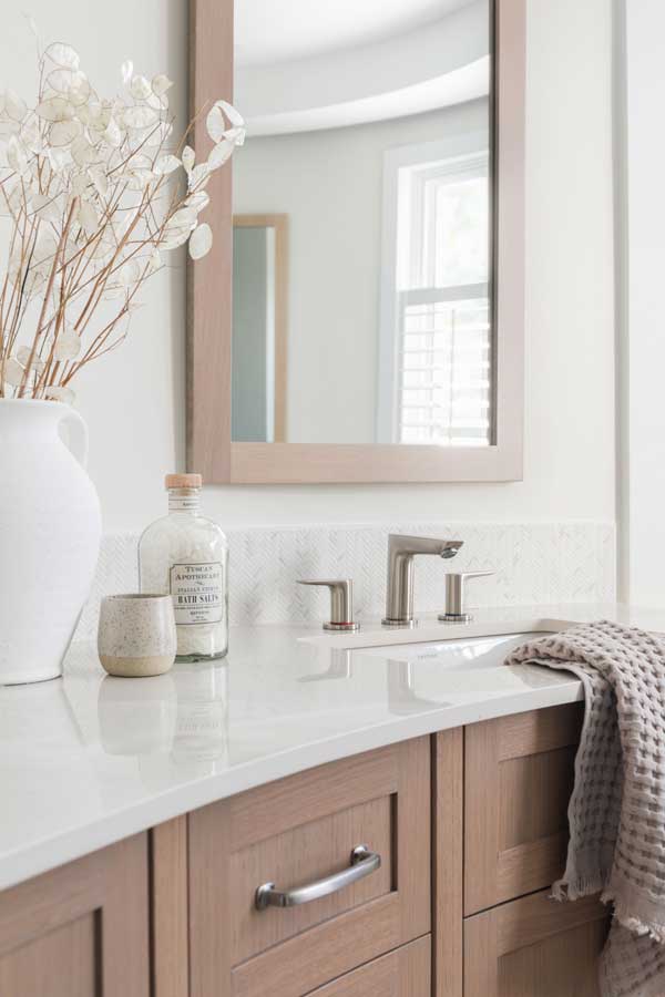 StudioRuInterior-Design_Calgary_Mount-Royal_Bathroom-Curved_Vanity This bathroom features a beautifully designed double vanity with warm wood cabinetry, soft neutral tones, and elegant detailing. The curved design maximizes space while adding visual interest. The light-colored stone countertop and herringbone-patterned tile flooring contribute to the room's sophisticated yet inviting ambiance. Large mirrors with wooden frames complement the cabinetry, reflecting natural light from the nearby window with plantation shutters. Thoughtful decor elements, such as the ceramic vase with delicate dried flowers and the textured hand towel, add a touch of warmth and character to this refined space.