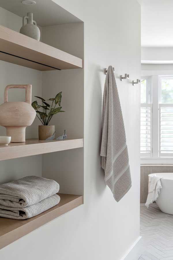 StudioRuInterior-Design_Calgary_Mount-Royal_Bathroom-Oak_Shelves This space exudes a serene and minimalist aesthetic with its clean lines, neutral tones, and thoughtfully curated decor. The built-in shelving with light wood finish offers both functionality and style, displaying soft, textured towels alongside elegant decorative pieces like ceramics and small sculptures. The hanging towel on the sleek metal hooks complements the soft color palette, enhancing the room’s spa-like feel. In the background, the freestanding tub and plantation shutters allow natural light to filter in, contributing to the overall calm and airy ambiance of the space.