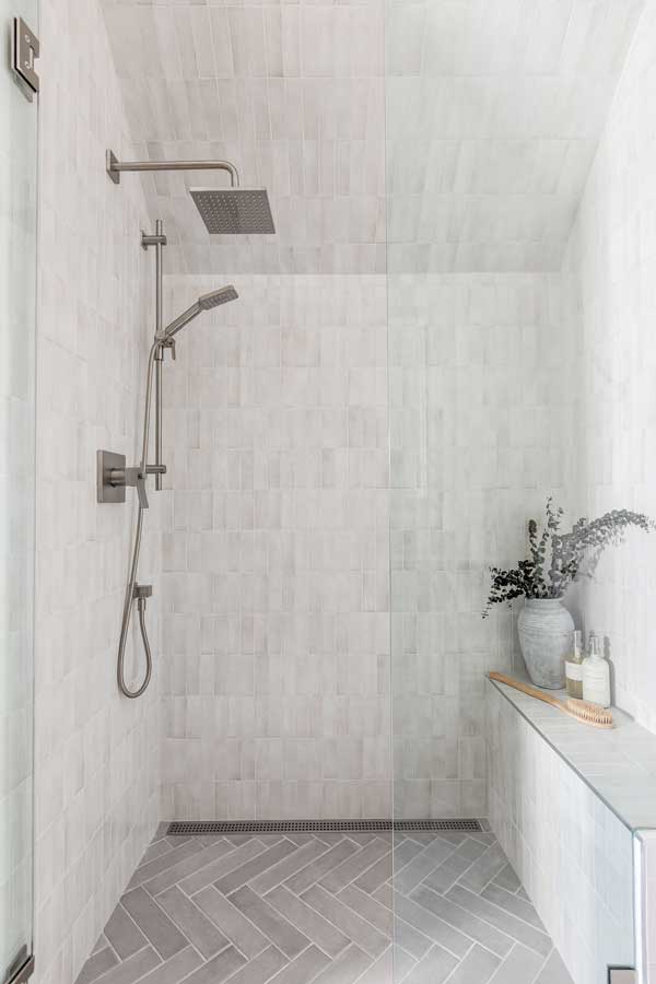 StudioRuInterior-Design_Calgary_Mount-Royal_Bathroom-Tiled_Shower This image showcases the beautifully renovated shower space. The transformation features a calming, neutral-toned aesthetic with soft, textured tiles covering the walls and ceiling. The large rainfall showerhead and sleek handheld fixture add a spa-like luxury, while the linear drain ensures a seamless and modern finish. The built-in bench, adorned with a ceramic vase and bath essentials, enhances both function and style. The herringbone-patterned floor tiles bring subtle contrast and depth, making this shower a stunning upgrade from its previous design.