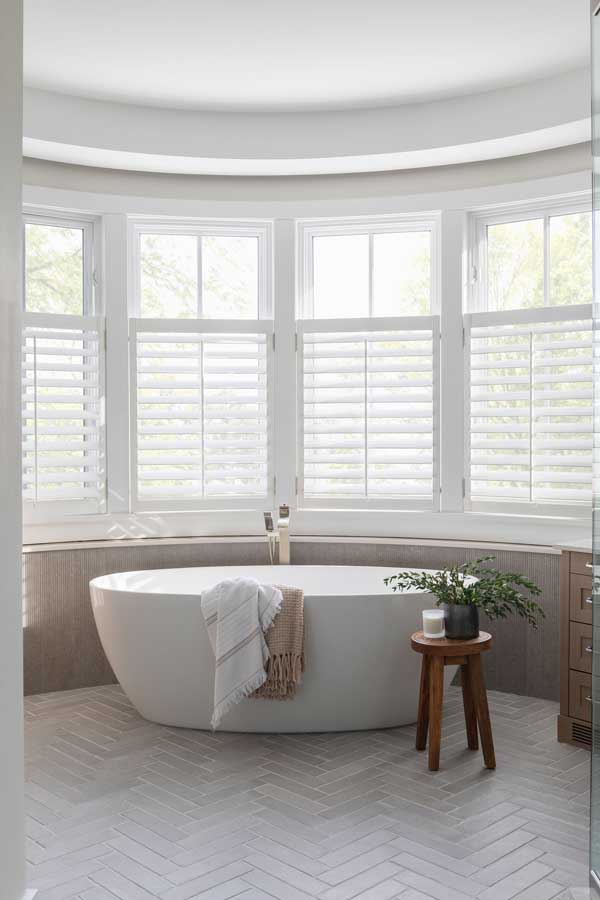 StudioRuInterior-Design_Calgary_Mount-Royal_Bathroom-Tub This bathroom exudes elegance and relaxation with its freestanding soaking tub placed in front of a curved wall of large windows, fitted with white plantation shutters that allow natural light to filter through. The combination of neutral tones, herringbone-patterned flooring, and textured wall paneling adds depth and sophistication. A wooden stool holding a potted green plant and a candle adds warmth, while draped towels over the tub enhance the inviting, spa-like atmosphere. The sleek floor-mounted faucet completes the modern yet timeless aesthetic.