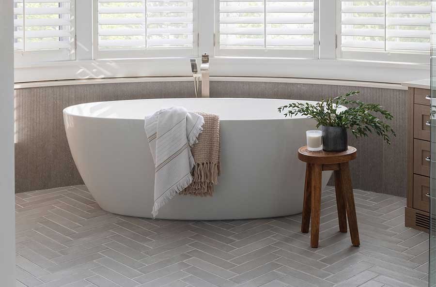 StudioRuInterior-Design_Calgary_Mount-Royal_Bathroom-Tub_Photo This bathroom exudes elegance and relaxation with its freestanding soaking tub placed in front of a curved wall of large windows, fitted with white plantation shutters that allow natural light to filter through. The combination of neutral tones, herringbone-patterned flooring, and textured wall paneling adds depth and sophistication. A wooden stool holding a potted green plant and a candle adds warmth, while draped towels over the tub enhance the inviting, spa-like atmosphere. The sleek floor-mounted faucet completes the modern yet timeless aesthetic.