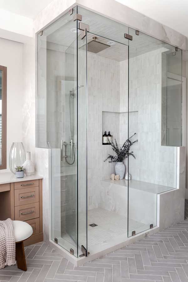 StudioRuInterior Design_Calgary_Mount Royal_Bathroom This bathroom exudes elegance and relaxation with its freestanding soaking tub placed in front of a curved wall of large windows, fitted with white plantation shutters that allow natural light to filter through. The combination of neutral tones, herringbone-patterned flooring, and textured wall paneling adds depth and sophistication. A wooden stool holding a potted green plant and a candle adds warmth, while draped towels over the tub enhance the inviting, spa-like atmosphere. The sleek floor-mounted faucet completes the modern yet timeless aesthetic.