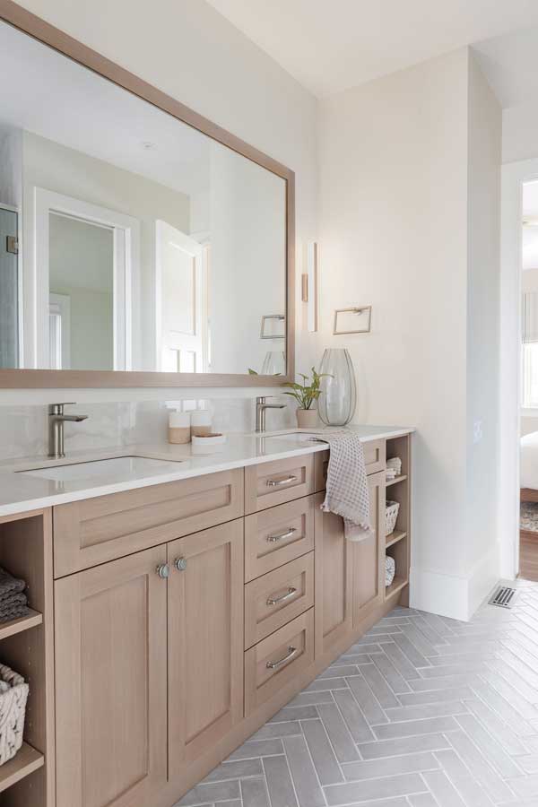 StudioRuInterior-Design_Calgary_Mount-Royal_Bathroom_Vanity This bathroom renovation features a warm and inviting double vanity with light wood cabinetry, sleek brushed nickel hardware, and a bright white quartz countertop. The large framed mirror enhances the sense of space, reflecting natural light from the adjacent bedroom. Built-in open shelving provides convenient storage for towels and essentials, while soft textures and neutral tones create a spa-like ambiance. The herringbone tile flooring adds a subtle yet sophisticated touch, seamlessly connecting the space with the rest of the bathroom. This transformation beautifully balances functionality and modern elegance.