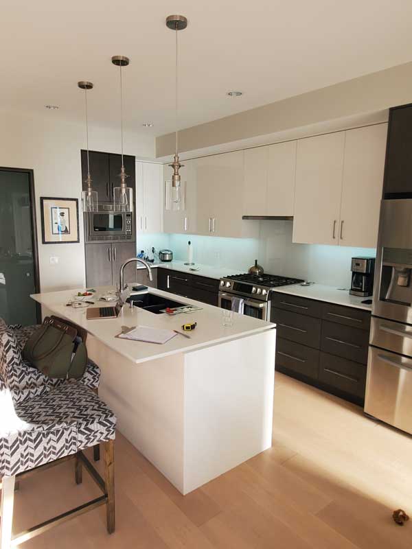 A modern kitchen with a clean and stylish design featuring a large white island with a waterfall edge, a stainless steel sink, and a high-arched faucet. The cabinetry is a combination of dark lower cabinets and light upper cabinets, providing contrast. Stainless steel appliances, including a refrigerator, oven, and microwave, are integrated into the space. Pendant lights with glass shades hang above the island, adding elegance. Light wood flooring brings warmth, while patterned barstools with wooden legs provide seating. Items such as a laptop, notebook, and measuring tape on the island suggest an ongoing project or workspace.