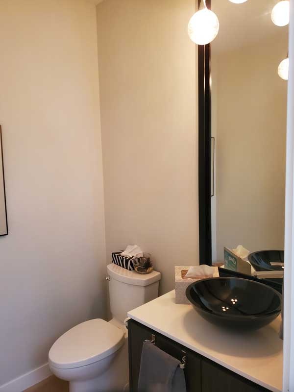 A modern powder room featuring a sleek black vessel sink atop a white countertop with dark cabinetry. A large mirror with a black frame extends vertically, reflecting the room’s lighting and adding depth. A contemporary toilet sits against the white walls, with decorative items such as a striped tissue box and a small glass container placed on top. Round glass pendant lights provide a warm glow, enhancing the clean and minimalist aesthetic. A towel hangs neatly on the vanity, completing the refined yet functional design.