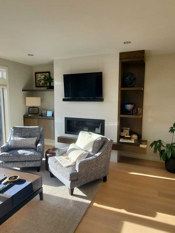 A modern kitchen with a sleek design featuring a large white island with a waterfall edge, dark lower cabinets, and light upper cabinets. Pendant lights with glass shades hang above the island, providing stylish illumination. Stainless steel appliances, including a refrigerator, oven, and microwave, complement the contemporary aesthetic. The light wood flooring adds warmth to the space, while patterned barstools with wooden legs provide seating. Various items, such as a laptop, notepad, and measuring tape, are scattered on the island, suggesting an active workspace.