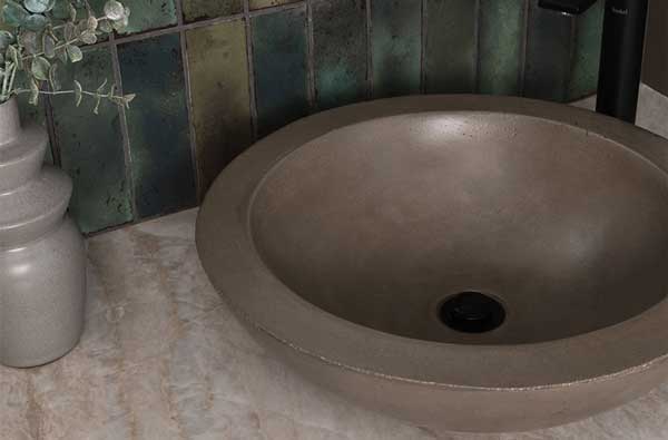 A modern bathroom vanity featuring a round, stone-like vessel sink with a matte finish, a sleek black faucet, and a beige marble-look countertop. The backsplash consists of rustic green-toned square tiles with an aged, weathered appearance. A curved mirror with a thin black frame is partially visible, reflecting a bright doorway. A decorative gray vase with dried eucalyptus adds a natural touch to the design.