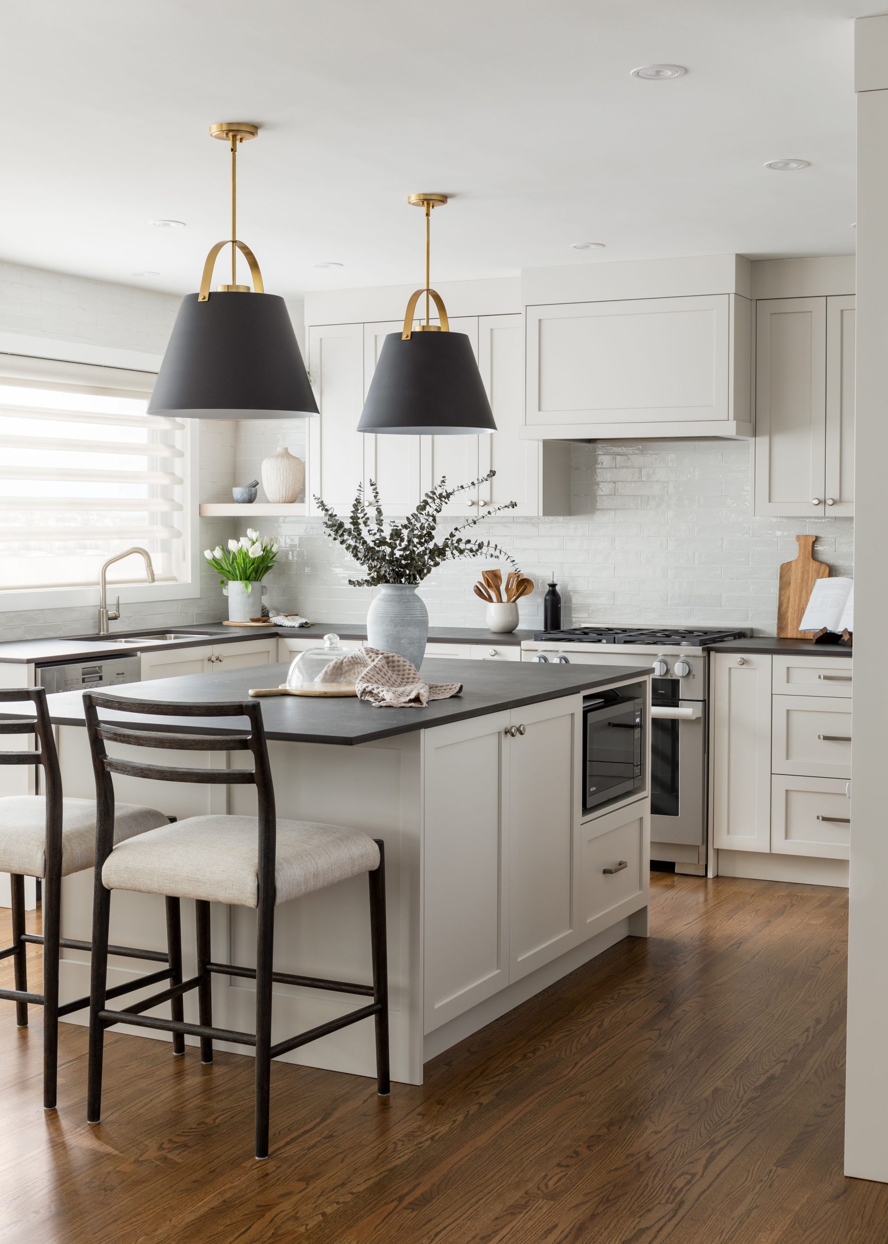 Modern kitchen with white cabinets, black pendant lights, and a central island featuring black and white bar stools. A vase with green foliage is on the counter.