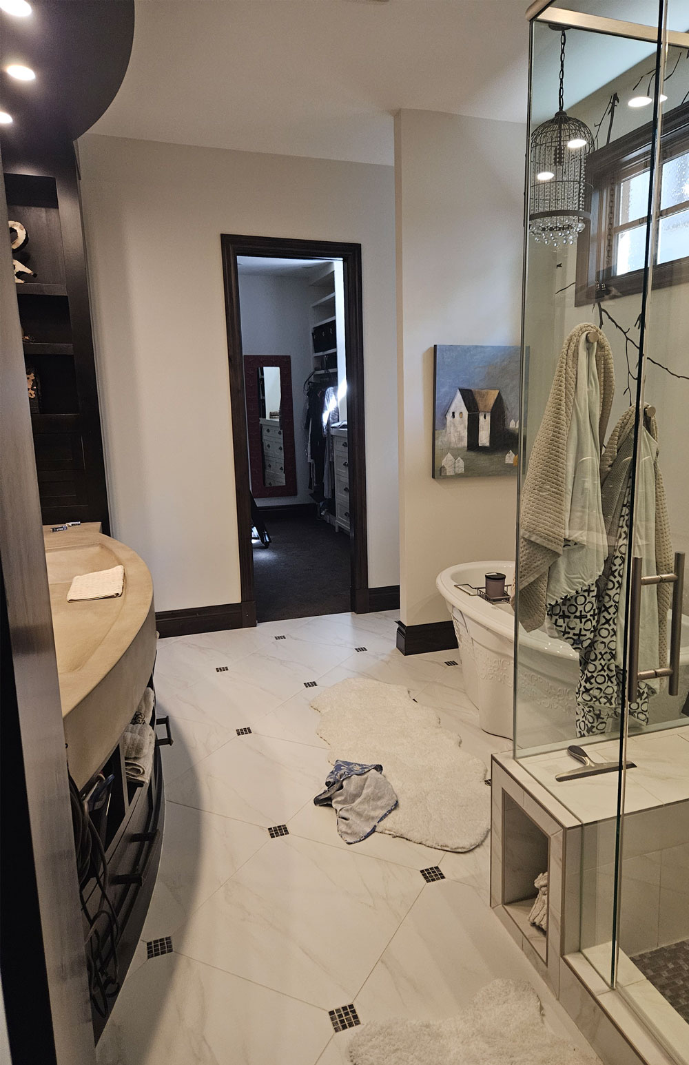 Before renovation: industrial-style ensuite with dark wood accents, curved concrete-look vanity, freestanding tub, glass shower and white tile flooring with dark insets.